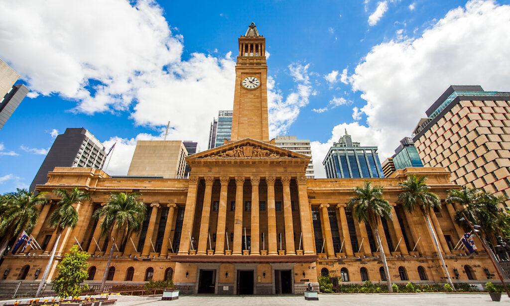 A grand building with a clock tower stands amidst skyscrapers, framed by palm trees. The clear sky and surrounding urban architecture create a vibrant cityscape.