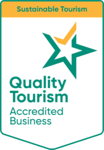 Badge featuring a green and gold star, labeled "Quality Tourism Accredited Business," on a white background. Above, an orange banner reads "Sustainable Tourism."